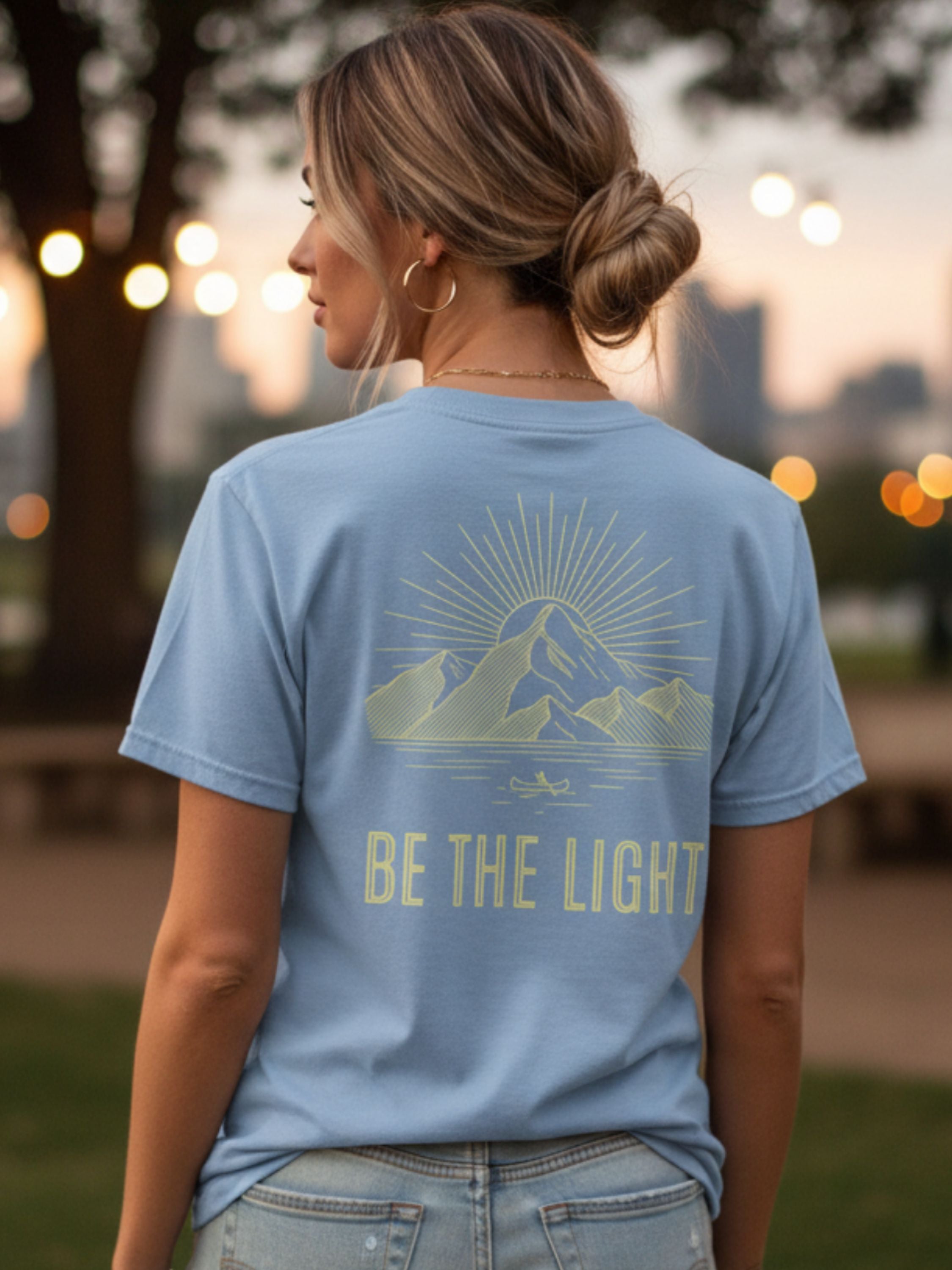 Person wearing a blue t-shirt with a mountain design and 'Be the Light' text, standing outdoors.