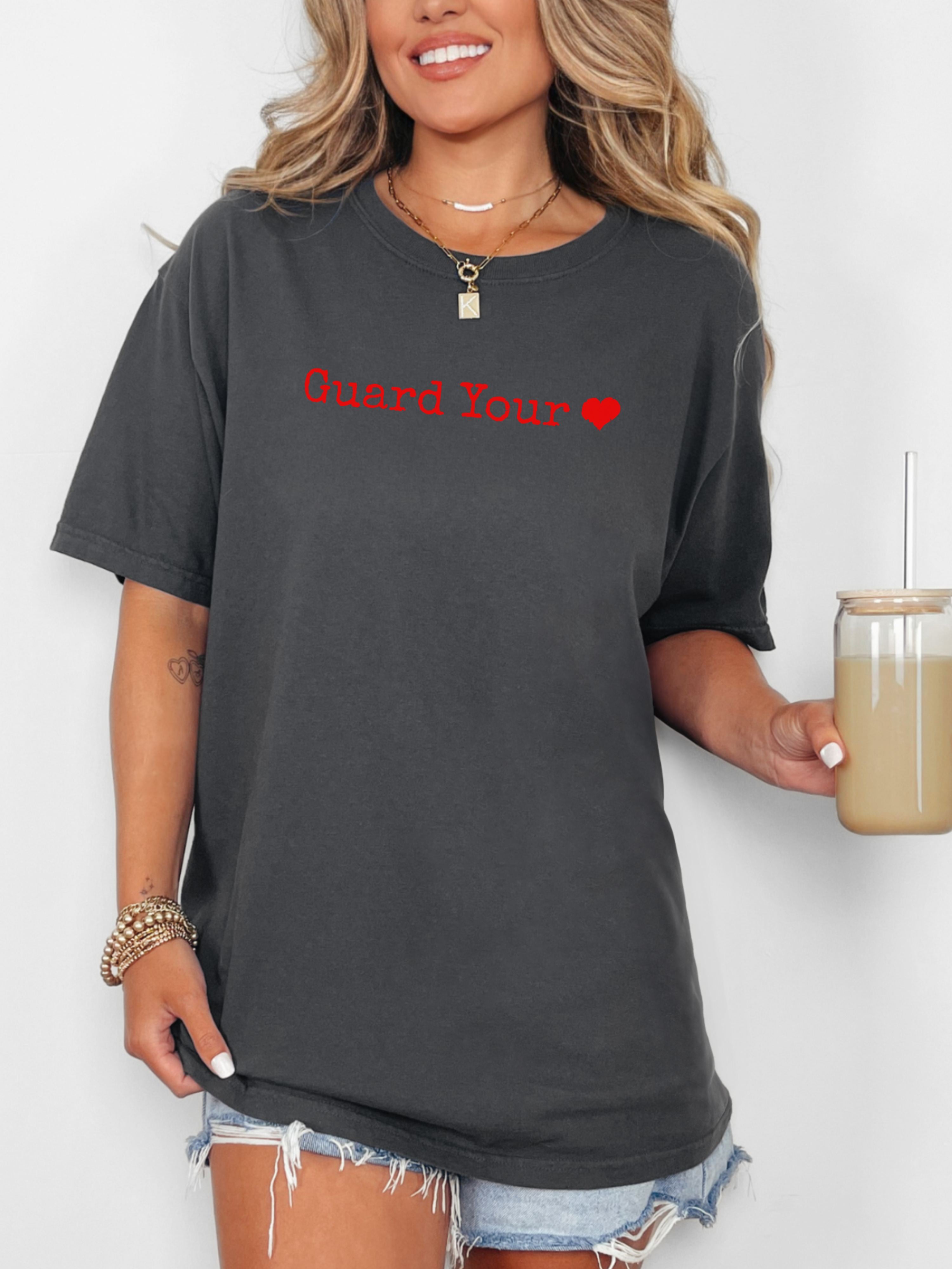 Woman wearing a dark gray t-shirt with 'Guard Your Heart' in red text, holding a drink.