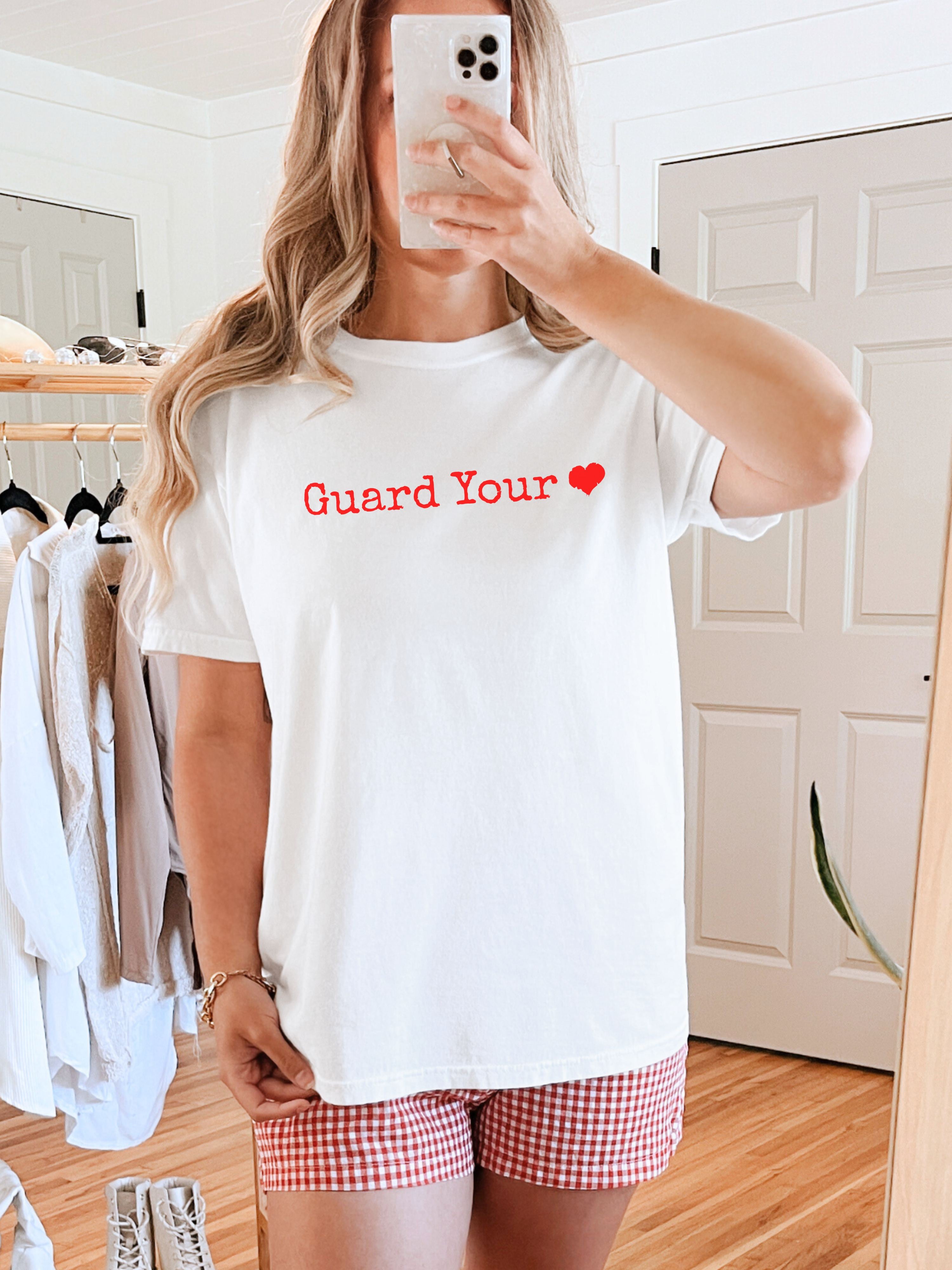 Woman wearing a white t-shirt with 'Guard Your Heart' red text in a room.