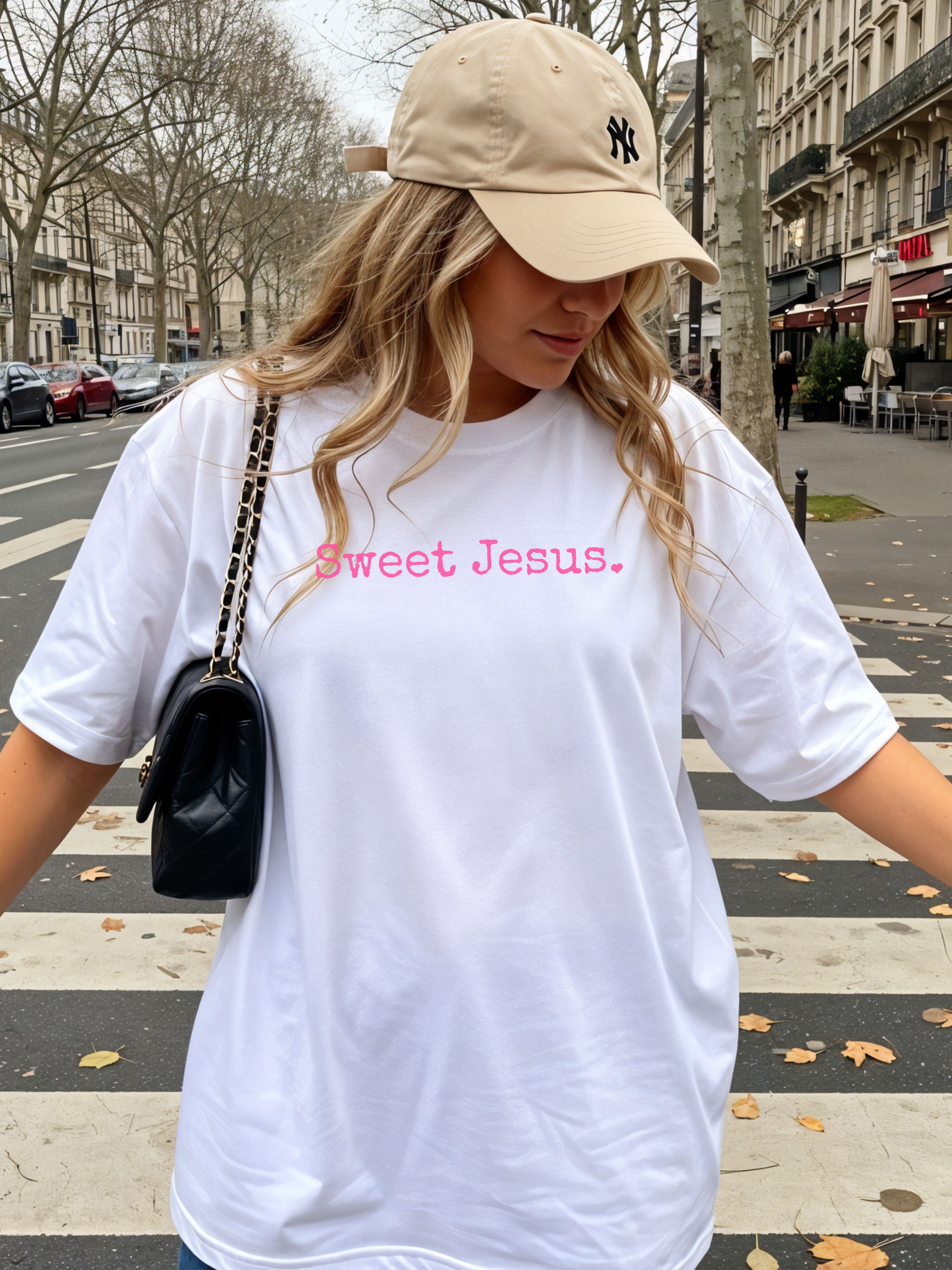 Woman wearing a white t-shirt with 'Sweet Jesus' text and a beige cap in an urban setting.