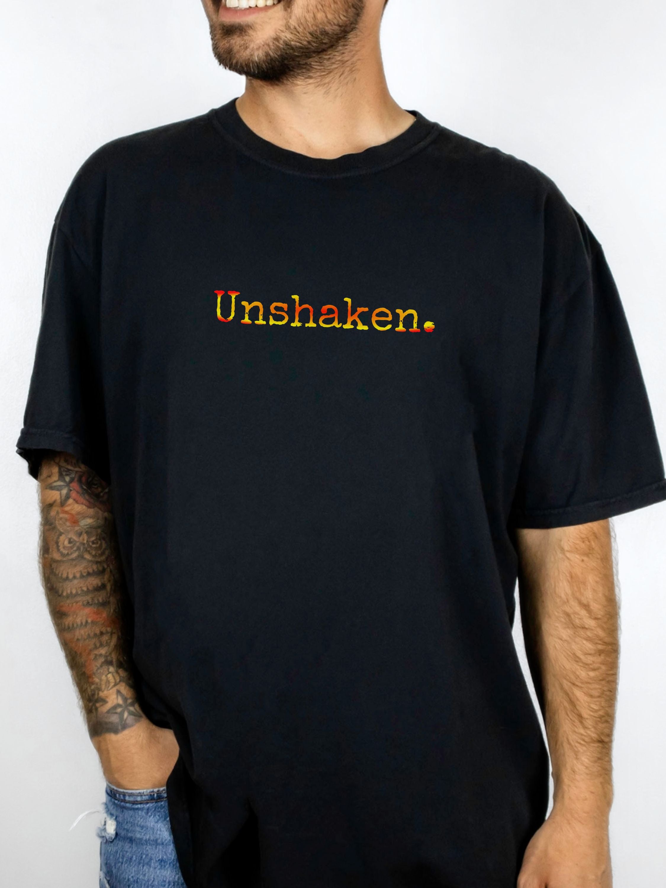 Male wearing a black t-shirt with 'Unshaken.' in red, orange and yellow text.