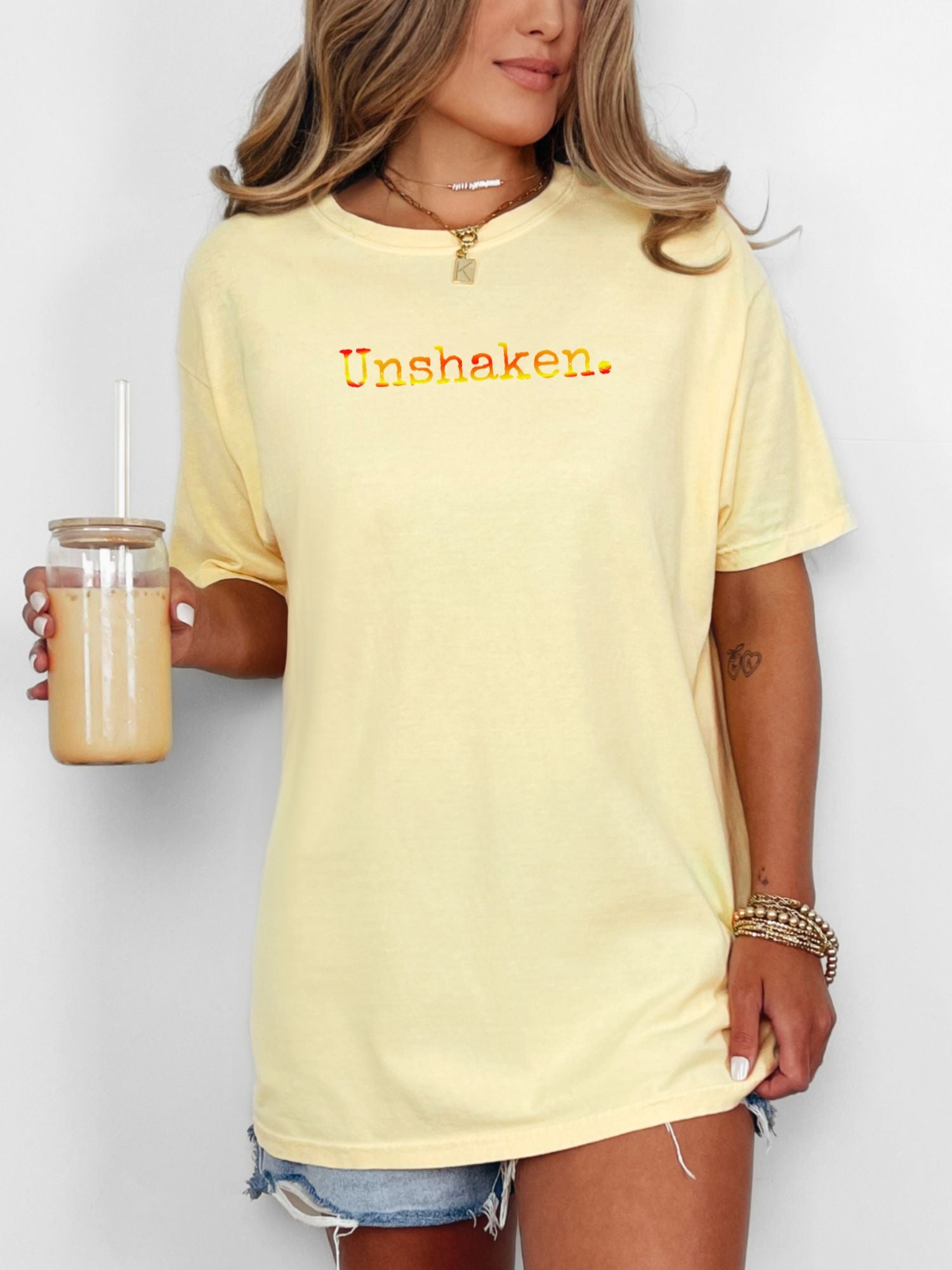 Female wearing a butter yellow t-shirt with 'Unshaken.' red, orange and yellow text.